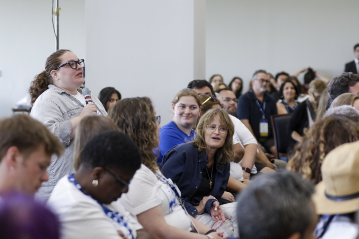 "Sarah Lerner speaks into a microphone during a packed session at the AFT TEACH 2025 conference. She stands among seated attendees, engaging with the panel while others listen attentively